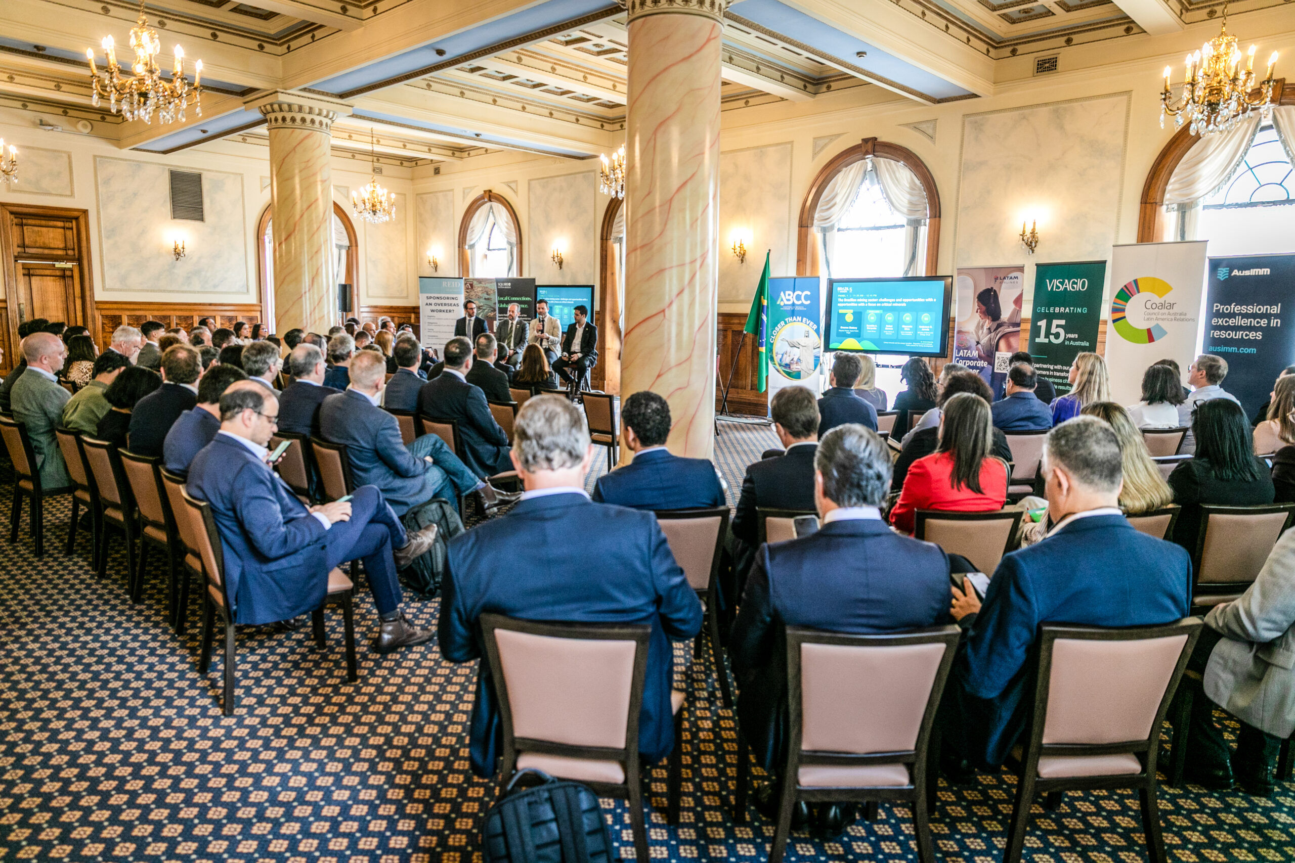 Audience seated in the event room, with screens and banners in the background.