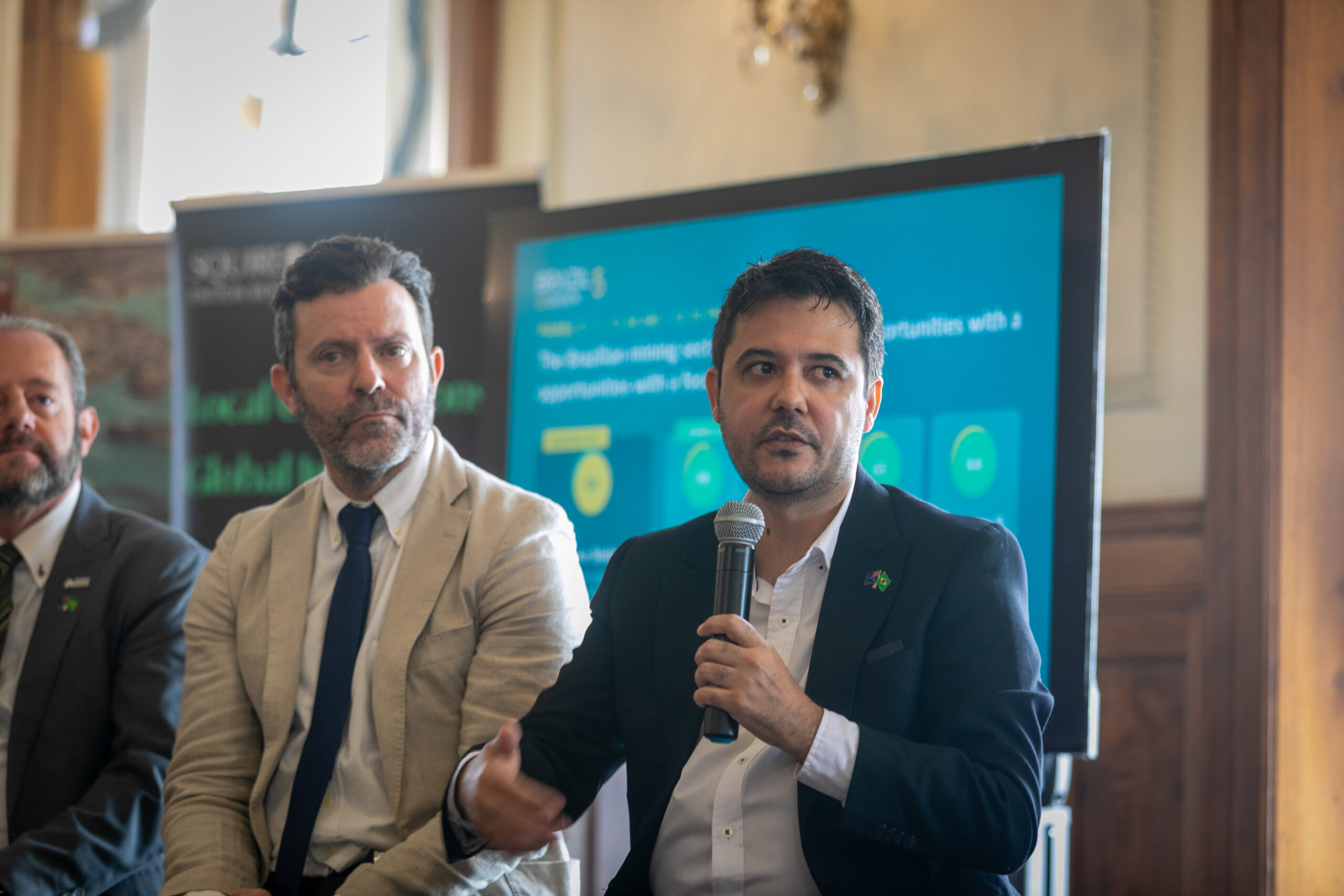 A panelist speaking into a microphone next to another panelist, with the panel slide in the background.